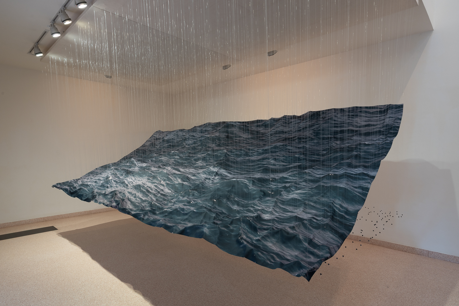 Une surface photographique figurant la mer est suspendue par des fils dans l’exposition Undercurrents de Miguel Rothschild.