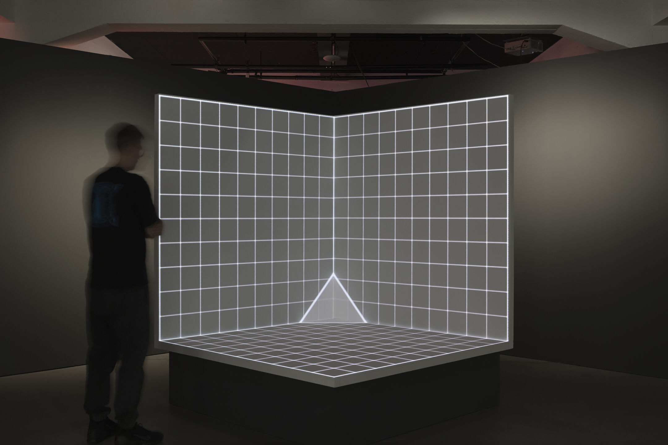 Projection d’une grille lumineuse blanche sur une structure angulaire, avec une silhouette de visiteur, dans Thresholds de Studio Lemercier au HEK.