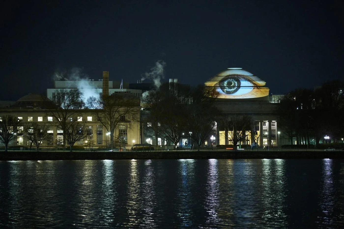 Un œil lumineux est projeté sur le Grand Dôme du MIT, vu de nuit depuis l’autre rive du Charles.