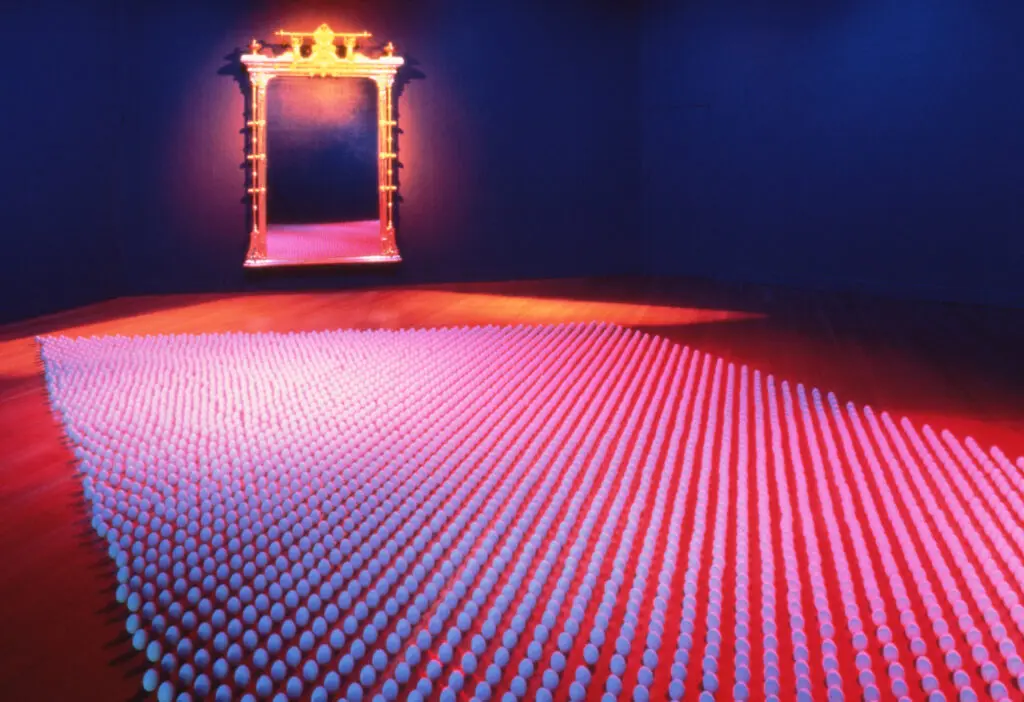 Installation Crossings de Mildred Howard : un champ d’œufs en céramique sous une lumière rouge et bleue, avec un cadre doré au fond