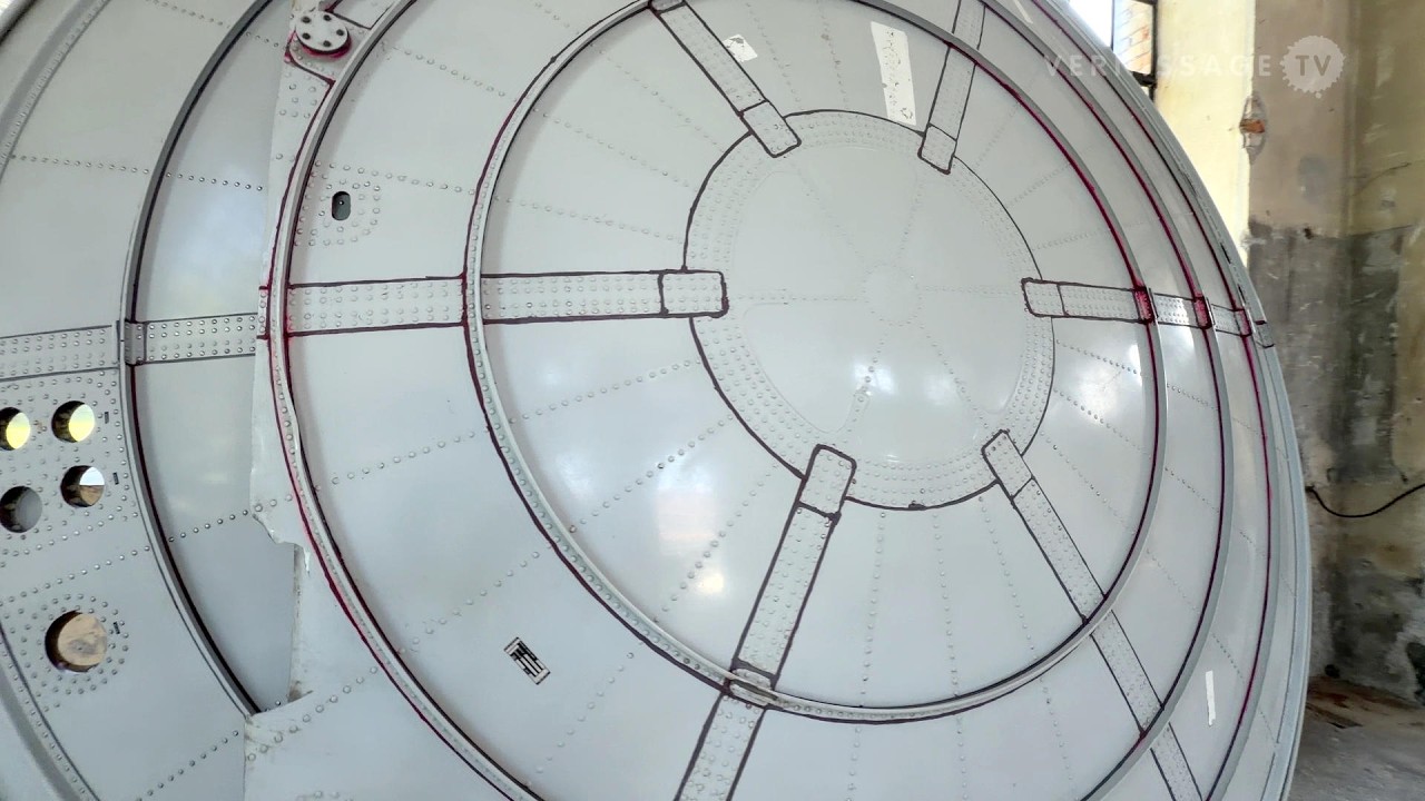 Grand élément circulaire en métal argenté, marqué de lignes et de traces, dans l’exposition HYLE.