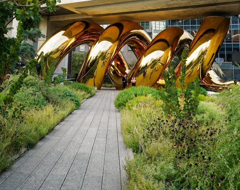 Une spirale gonflable dorée et réfléchissante traverse la promenade plantée de la High Line, sous un bâtiment, devant deux visiteurs.