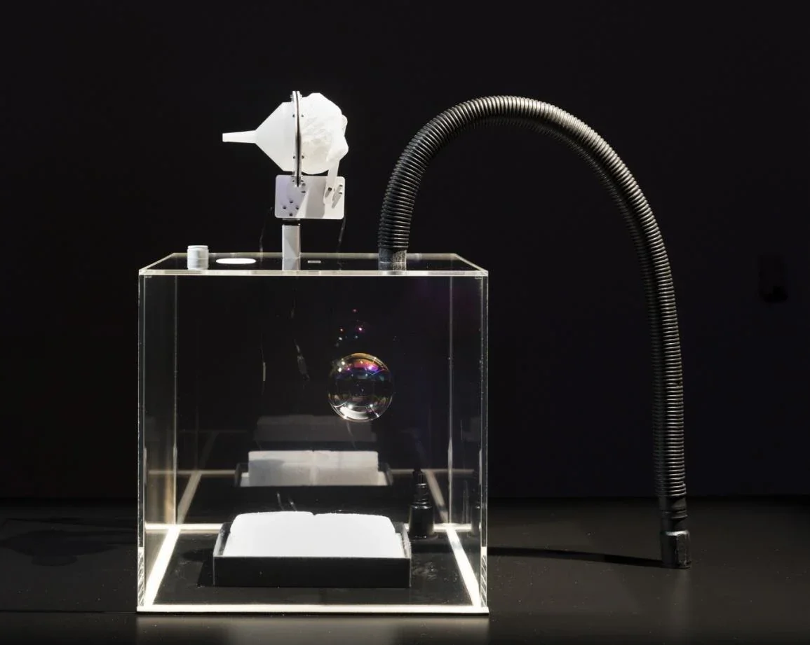 Une bulle de savon flotte dans un cube transparent relié à un dispositif mécanique et à un tuyau noir.