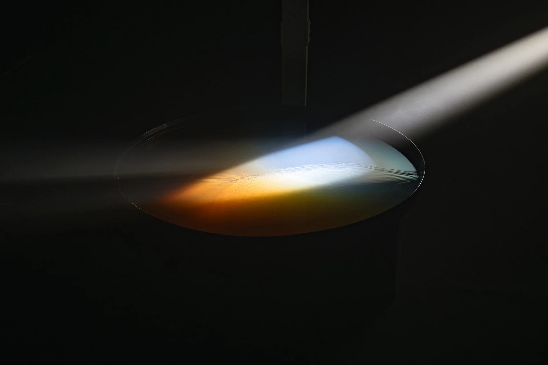 Un faisceau blanc frappe un miroir circulaire couvert d'eau, traversé de reflets bleus et orangés dans l'obscurité.