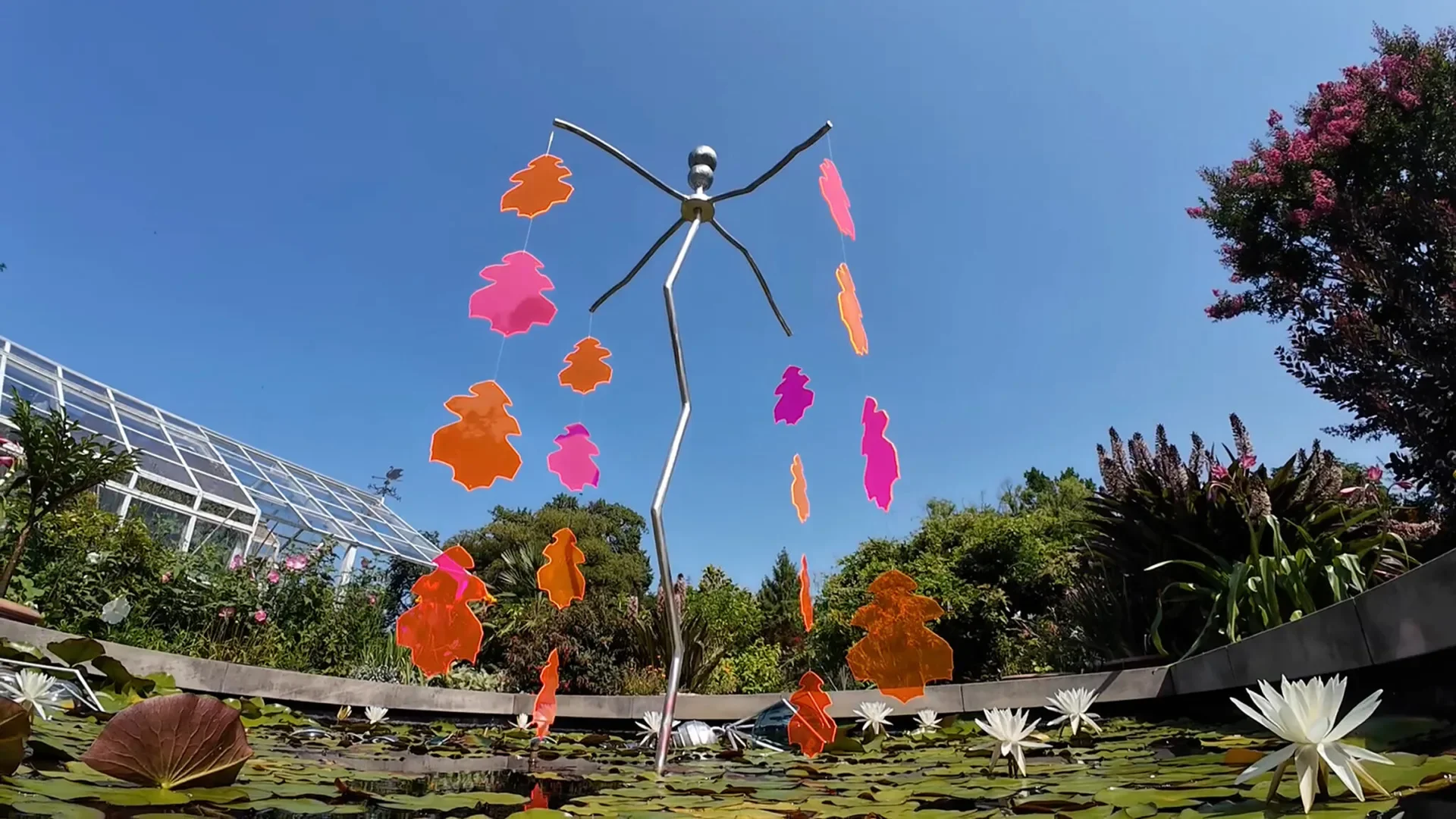 Une sculpture métallique flottante porte des formes roses et orange au-dessus d’un bassin de nénuphars.