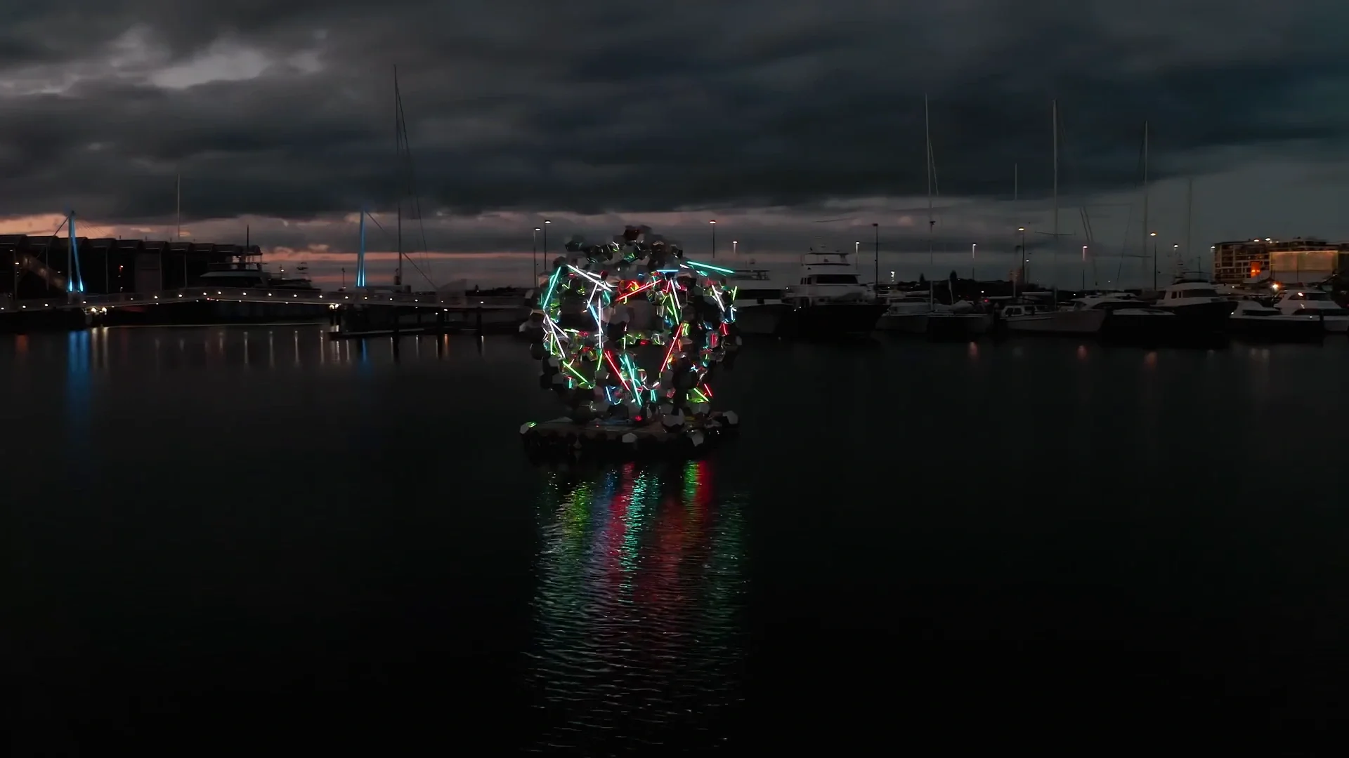 Une sculpture géométrique d’acier réfléchissant et de néons colorés flotte sur un ponton dans le port d’Auckland au crépuscule, avec son reflet dans l’eau.