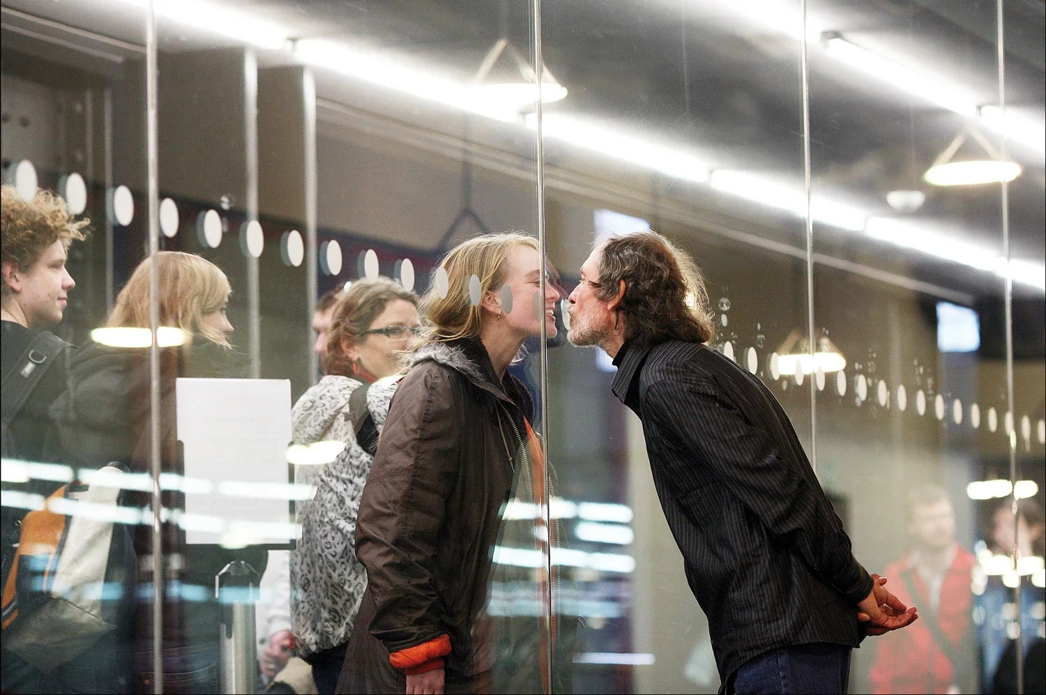 Jiří Kovanda et une participante se rapprochent pour s’embrasser de part et d’autre d’une paroi de verre à Tate Modern en 2007, devant plusieurs témoins.