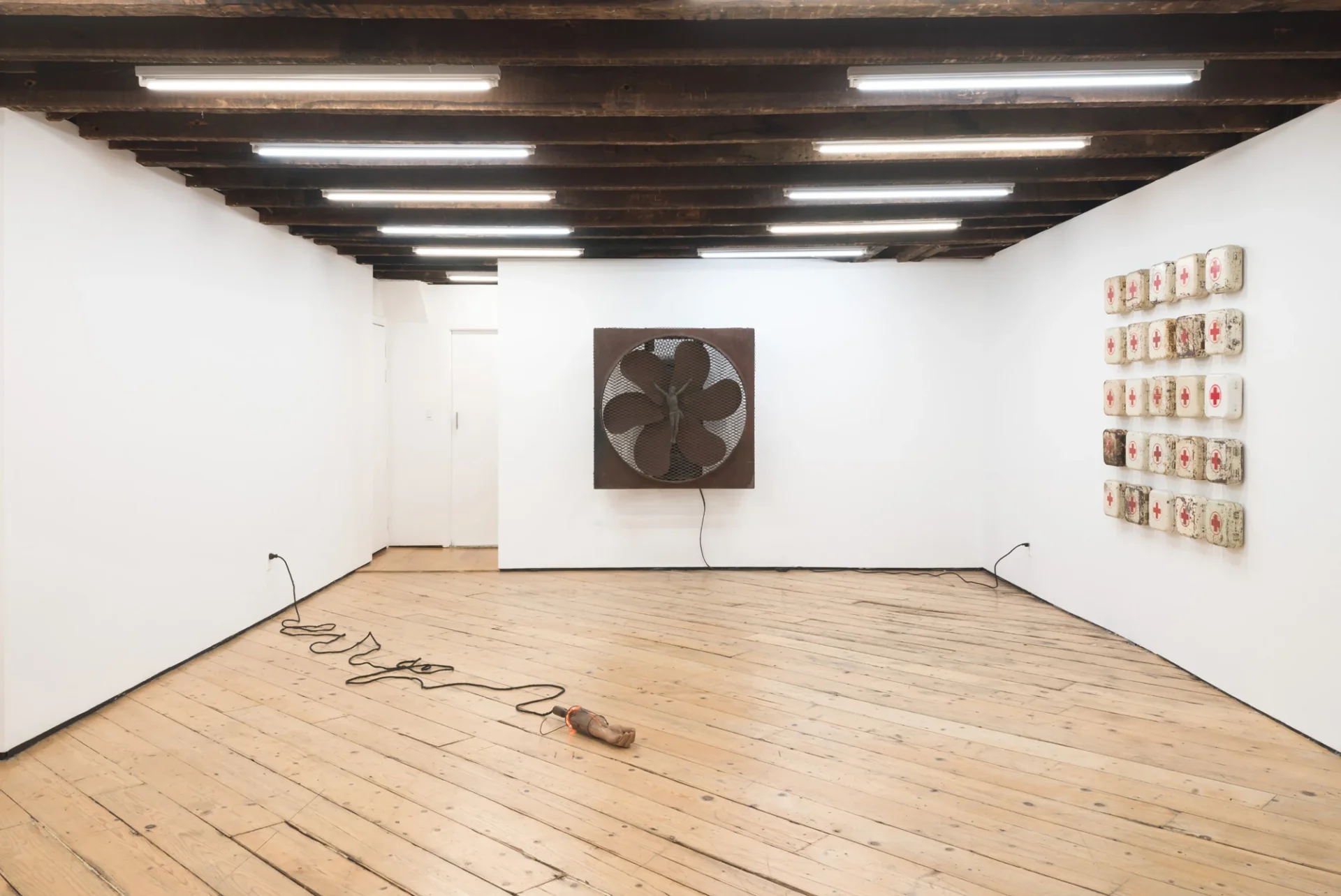 Vue de l’exposition de Jonah Dillon : ventilateur portant une figure du Christ, bras prothétique au sol et grille de trousses de secours.