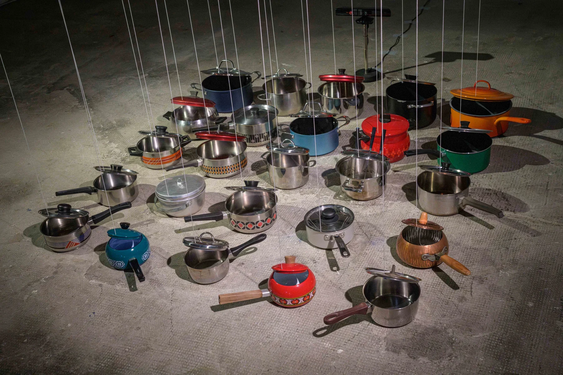 Vingt-cinq casseroles de couleurs et de formes variées, leurs couvercles reliés à des fils, dans l’installation de Michel Winterberg à E-WERK Freiburg.