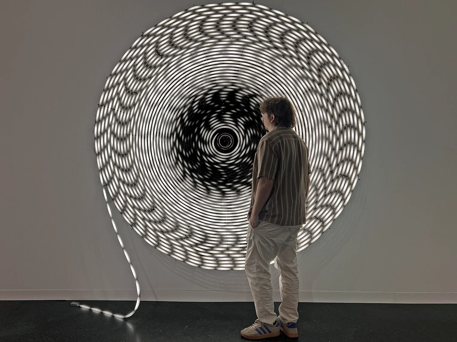 Un visiteur devant Black Hole de Rafael Lozano-Hemmer : une spirale lumineuse concave dessine son portrait autour d’une caméra centrale.