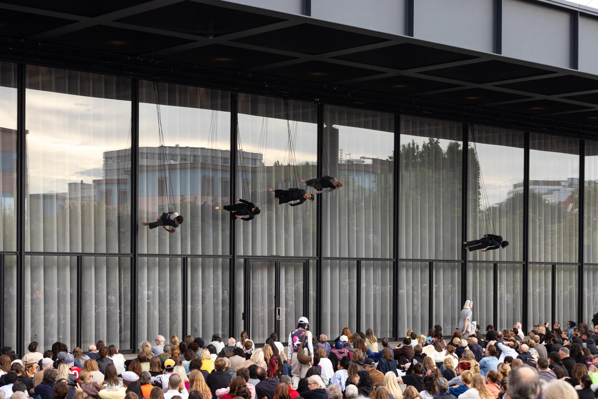 Interprètes suspendus horizontalement devant la façade vitrée de la Neue Nationalgalerie et le public.