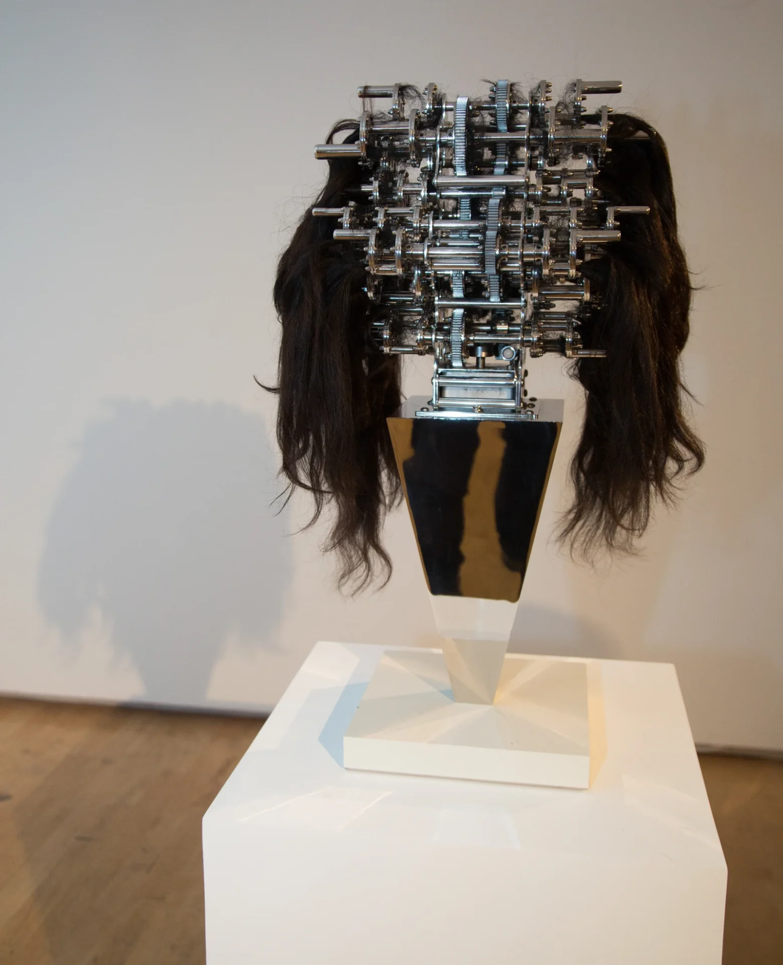 Une structure de tiges et d’engrenages chromés, entourée de longues mèches noires, sur un socle en miroir : Machine with Hair Caught in It d’UJOO+LIMHEEYOUNG.