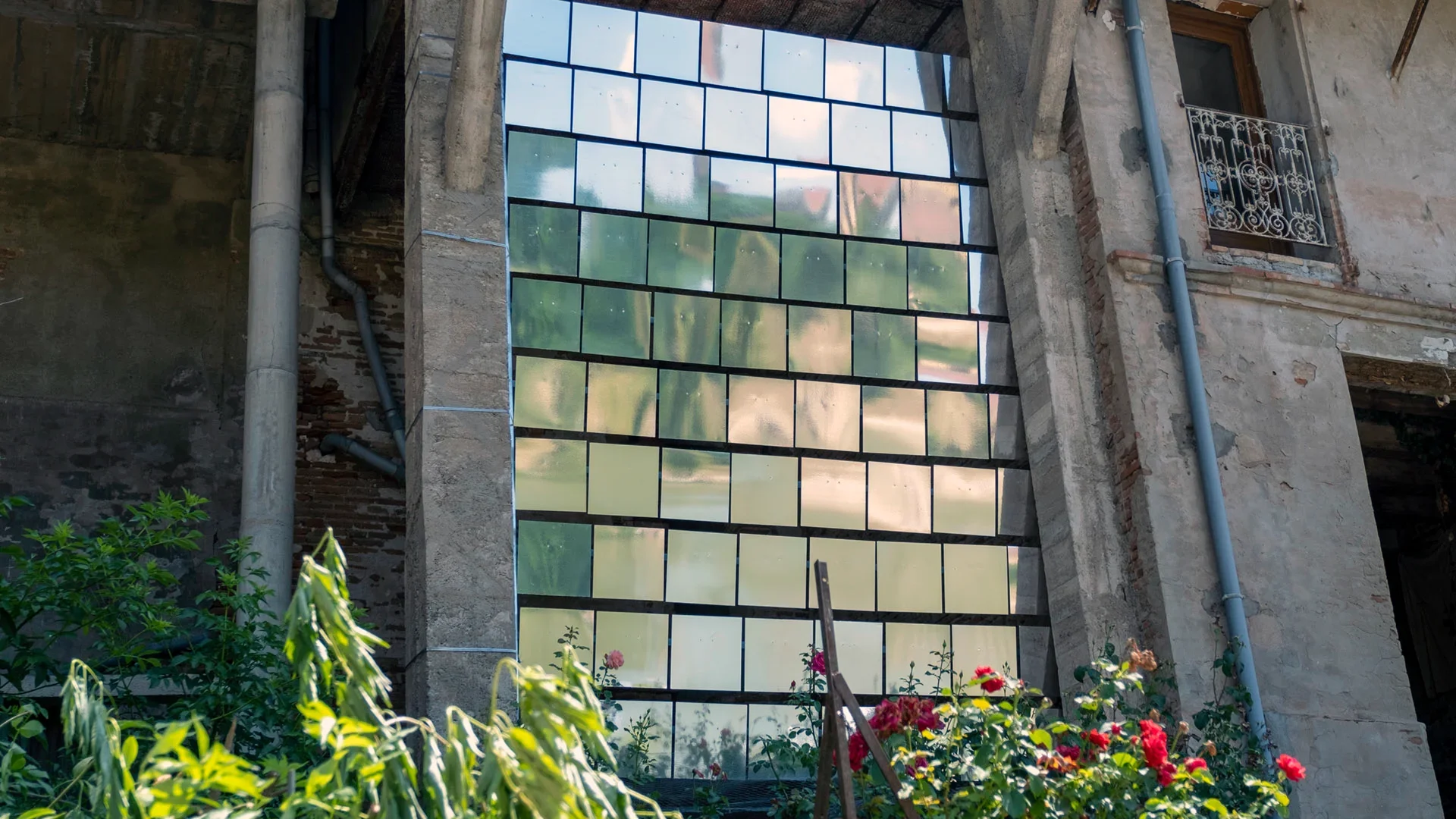 Écran de petits miroirs rectangulaires entre deux murs anciens, reflétant la lumière et la végétation.